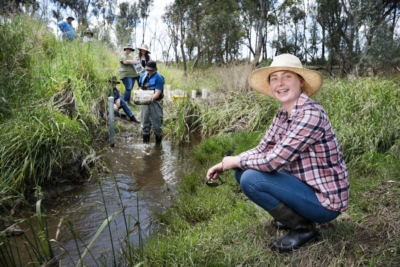 A woman wearing a straw hat kneels next to a stream where in the background man stands in the water examining the contents of a next while 5 other people look on