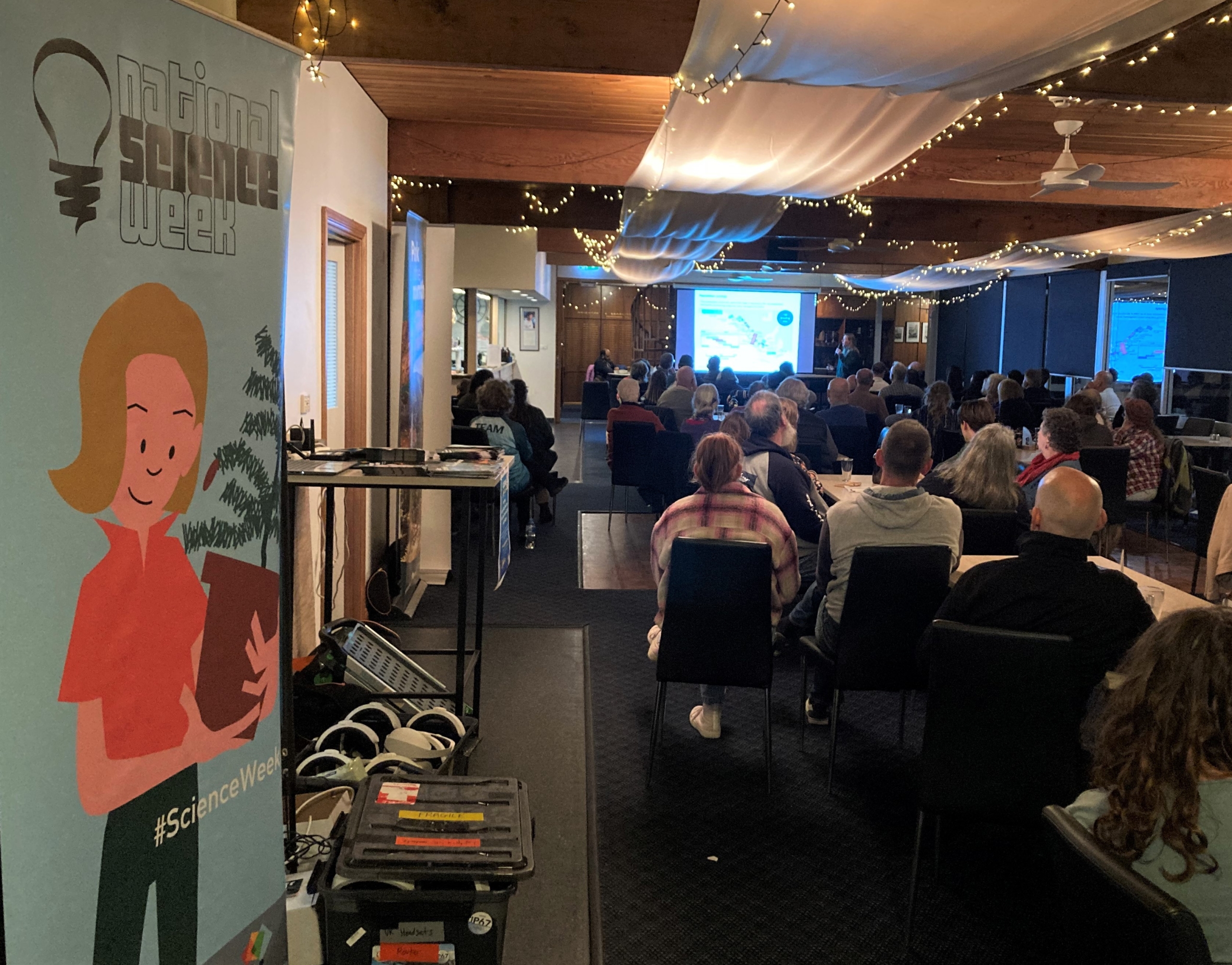 a photo taken from the back of the room with a National Science Week on the left. An audience sits in chairs facing a speaker who is speaking with a power point presentation on a screen 