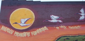 a section of a mural featuring white birds flying across the sun with Kaurna words underneath and the words "We Walk Together"